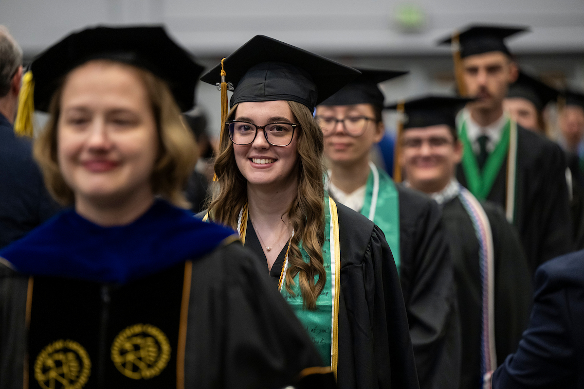 Students in graduation caps and gowns walk in a line, smiling. A woman in a faculty gown is in the foreground. The atmosphere is joyful and celebratory.